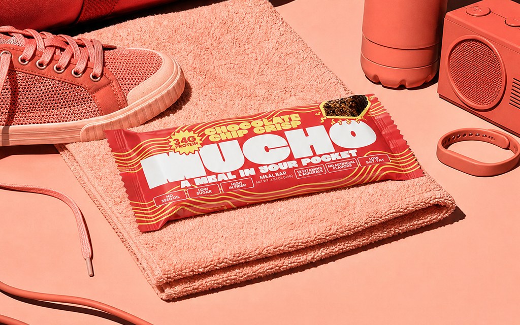A Mucho protein bar on a towel surrounded by gym kit — sneaker, water bottle, dumbbells and skip rope.